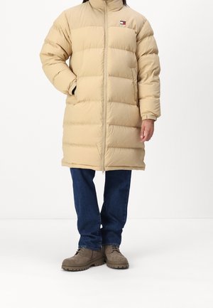 Personne portant un long manteau matelassé beige avec fermeture éclair, un jean bleu et des bottes marron, debout avec une main dans la poche du manteau.