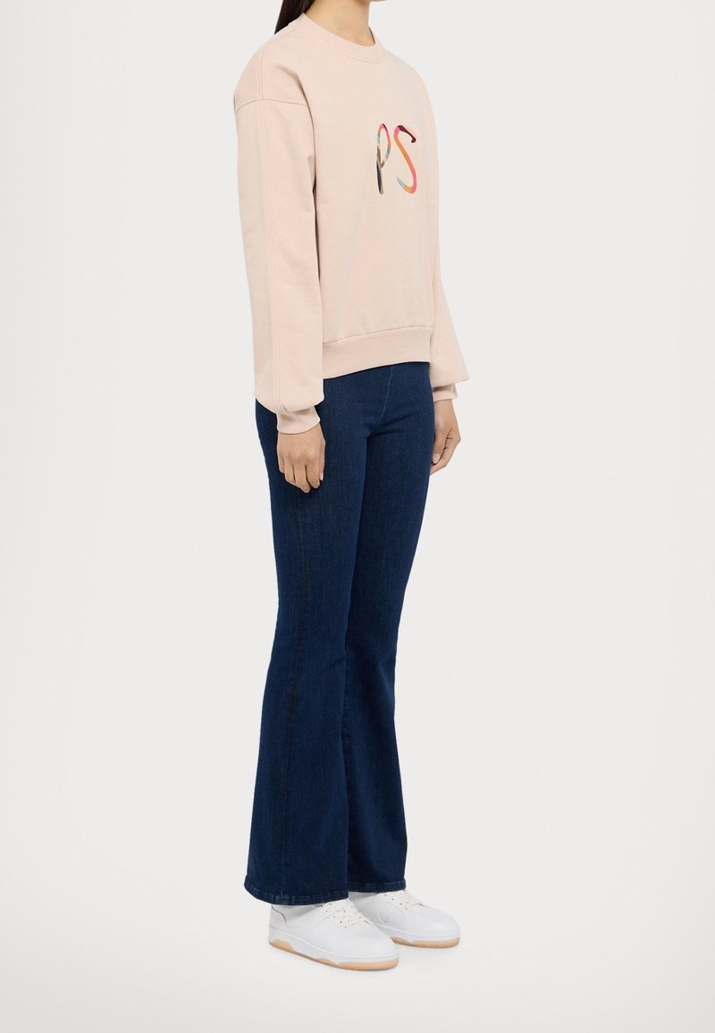Personne portant un sweat-shirt rose clair avec un logo multicolore "PS", un jean évasé bleu foncé et des baskets blanches, debout de profil.