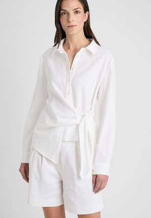 Femme portant une chemise blanche à manches longues avec col et nœud sur le côté, ainsi qu’un short blanc assorti à la hauteur du genou, debout devant un fond uni.