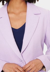Blazer in lana lavanda con colletto a punta, chiusura a un bottone e superficie morbida. Indossato sopra un top blu navy a scollo a V.