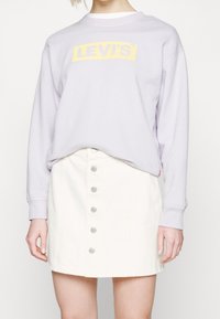 Jasnofioletowa bluza z żółtym logo "LEVI'S", w połączeniu z białą spódnicą z guzikami z przodu oraz srebrnymi guzikami i delikatnym rozkloszowaniem.