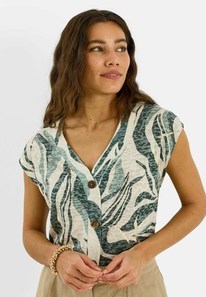 Femme aux longs cheveux ondulés portant un haut boutonné à manches courtes vert et blanc à motifs et un pantalon beige, mains jointes devant.