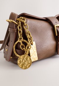 Bruine leren handtas met gouden ketting en twee getextureerde gouden charmes die aan de zijkant hangen, dichtbij de sluiting.