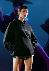 Čierna mikina s kapucňou s logom "HUNTRX" a pruhovanými rukávmi. Model má na sebe metalické šortky s hladkou textúrou a voľným strihom.