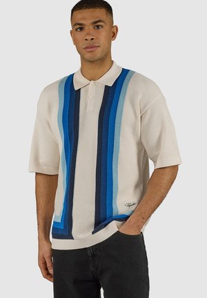 Mężczyzna w beżowym, dzianinowym polo w pionowe niebieskie paski i czarnych spodniach, stojący z jedną ręką w kieszeni na tle jednolitego tła.