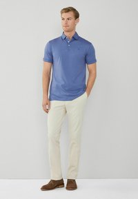 Uomo che indossa una polo blu a maniche corte, pantaloni beige e scarpe in suede marroni, in piedi con una mano in tasca su uno sfondo semplice.