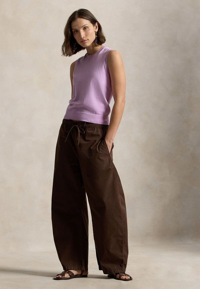 Γυναίκα που φοράει μωβ αμάνικη μπλούζα, παντελόνι σε σκούρο καφέ χρώμα με φαρδιά πλάγια και κορδόνι στη μέση, και καφέ σανδάλια, στέκεται μπροστά σε ουδέτερο φόντο.