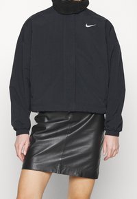 Svart Nike-jacka med en shearlingkrage, långa ärmar och en kort design. Kombinerad med en svart läderskjorta. Minimalistisk look.