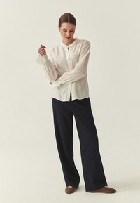 Blouse boutonnée beige clair avec manches longues froncées, associée à un pantalon large bleu marine foncé. La tenue présente une coupe décontractée et un design minimaliste.