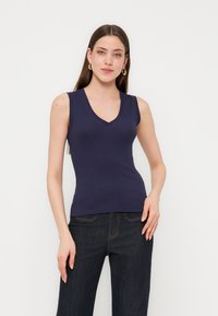 Jeune femme aux longs cheveux bruns portant des boucles d'oreilles créoles dorées, un haut bleu marine sans manches à col en V, et un jean foncé, posant devant un fond blanc.