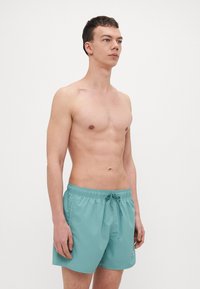 Turkoosi uimapuku, joka on valmistettu kevyestä materiaalista. Siinä on elastinen vyötärö, jossa on nyöri, sekä pieni logokoristelu sivussa.