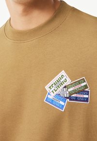 Hnědá mikina s kulatým výstřihem, zdobená barevnými vyšívanými nášivkami s nápisy "Tenis", "Golf" a "Sport" v různých barvách a obdélníkových tvarech.