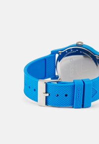 Modré silikónové hodinky s okrúhlym puzdrom, strieborným kovovým prevedením, textúrovaným remienkom a lešteným zadkom s gravírovaným logom značky.