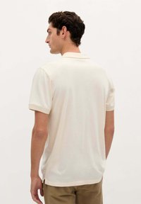 Camisa polo de color crema con mangas cortas, textura suave y cuello. Diseño liso con detalles de costura sutiles, combinada con pantalones khaki claros.