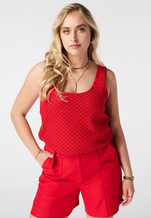 Vrouw met lang blond haar, gekleed in een rood mouwloos gehaakt topje en rode shorts, poserend met één hand in de zak tegen een witte achtergrond.