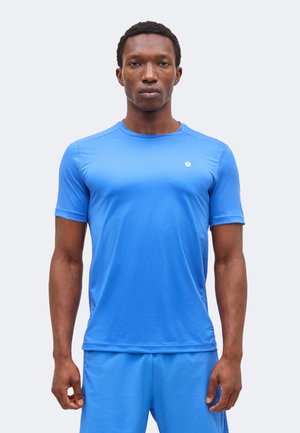 Mand stående fremadrettet iført en tætsiddende lyseblå kortærmet skjorte og matchende blå shorts mod en ensartet baggrund.