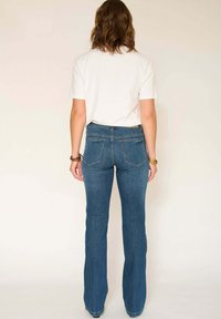 Femme aux cheveux bruns mi-longs portant un t-shirt blanc, un jean bootcut bleu et des bracelets, debout devant un fond beige uni.