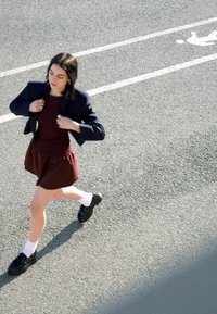 O persoană poartă un sacou închis la culoare peste o rochie burgundă cu fustă evazată. Pantofi negri asociați cu șosete albe. Fundalul este o șosea asfaltată cu marcaje.