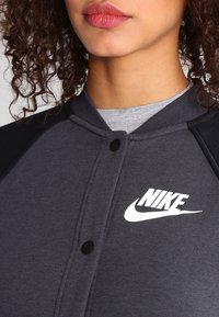 Grå polyesterjacka med svarta accenter, rund krage, knappstängning och broderad vit Nike-logga på vänster bröst.