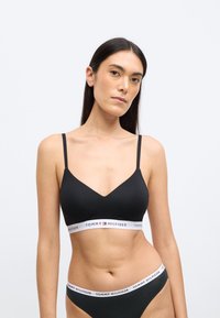 Črni bralet z tankimi naramnicami in V-izrezom. Ima elastični trak z znamko v beli barvi z rdečimi in modrimi poudarki. Gladek tekstura.