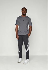 T-shirt de sport gris à manches courtes avec des accents noirs, associé à un pantalon sombre avec trois rayures blanches sur les côtés. Baskets blanches.