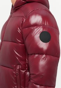 Section of shiny burgundy puffer jacket with side zipper pocket and black circular patch on upper arm.