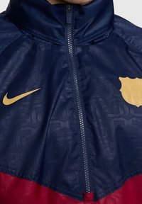 Chaqueta de Nike con una parte superior azul marino con un patrón texturizado y una sección inferior roja. El logotipo y los acentos en dorado son prominentes. Con cremallera en la parte delantera.