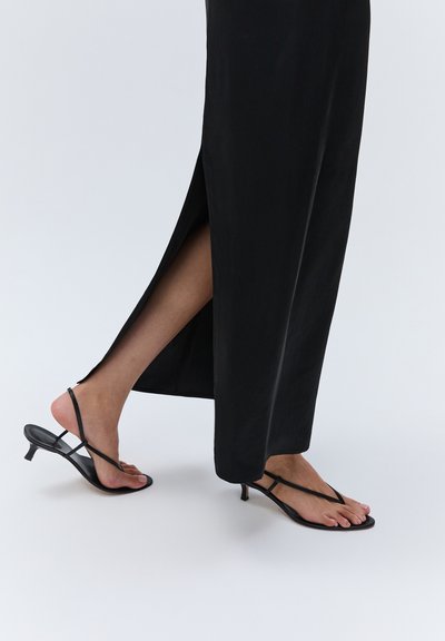 Personne portant des sandales noires à brides et petit talon ainsi qu'une longue jupe noire avec une fente latérale dévoilant une jambe nue.