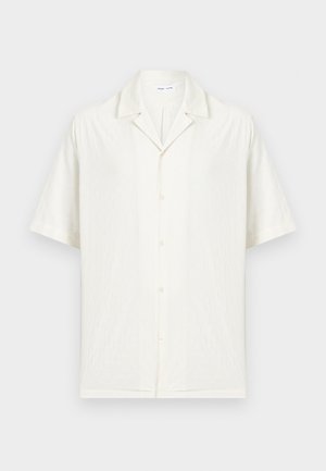 Chemise blanche à manches courtes avec col campagnard et tissu texturé subtil, présentée sur un fond clair uni.