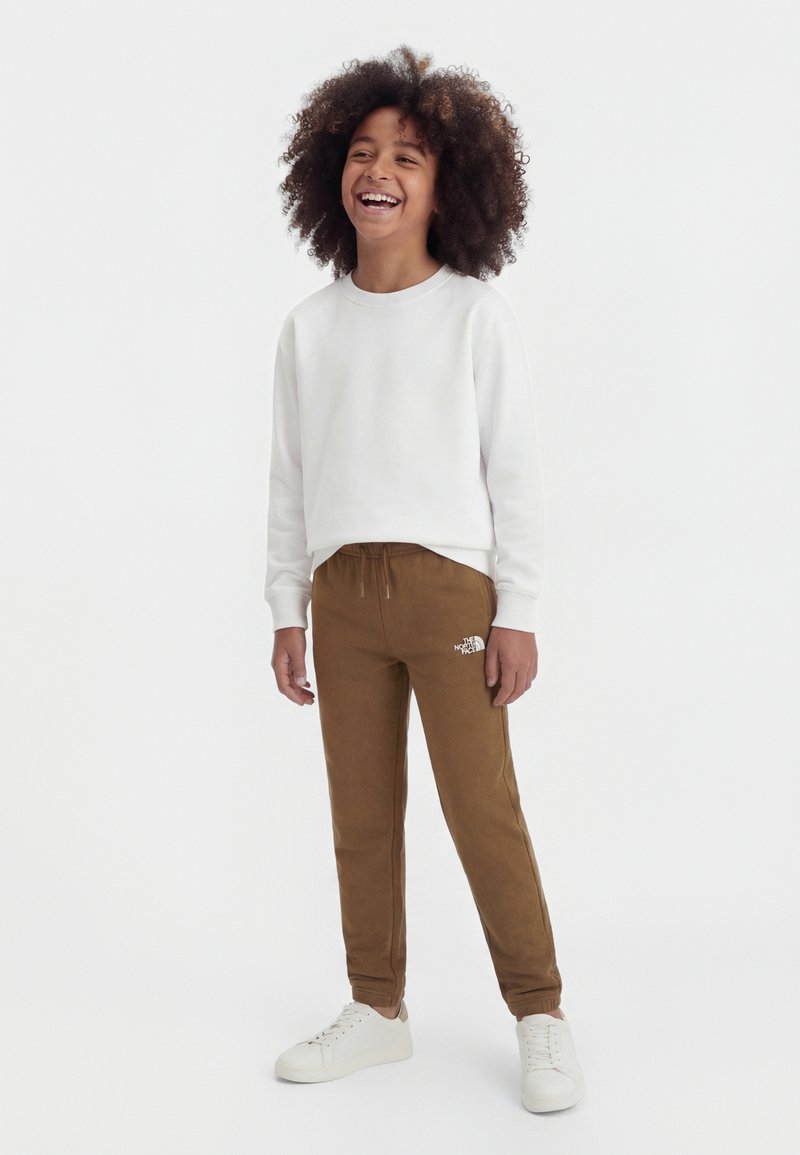 Barn med krøllet hår iført hvid sweatshirt, brune North Face joggingbukser og hvide sneakers, smilende og stående på hvid baggrund.