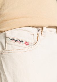 Béžové džíny z denimu s malou obdélníkovou nášivkou, která zobrazuje "Diesel Industry Denim Division" v červené a šedé barvě. Měkká textura, slim fit.