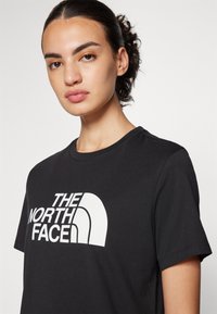 Must must värviline T-särk, millel on üle rinna suur valge "The North Face" logo. Lühikesed varrukad ja ümar kaelus.