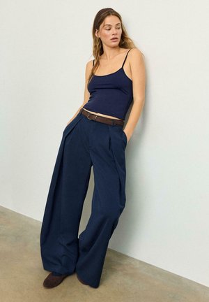 Donna con top a spalline sottili color blu navy, pantaloni a gamba larga blu navy con cintura marrone e scarpe marroni, in piedi contro un muro bianco.