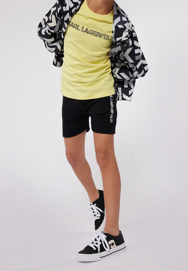 Schwarz-weiße gemusterte Jacke über einem gelben Grafik-T-Shirt, kombiniert mit schwarzen Shorts mit weißem Logo und schwarzen Schuhen mit einem Icon.