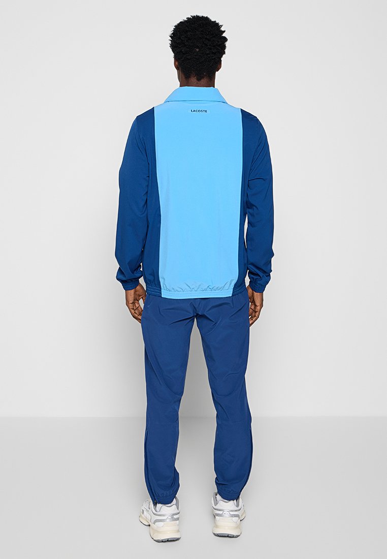 Uomo di spalle che indossa una giacca Lacoste blu a due toni e pantaloni blu abbinati, con sneakers bianche, posato su uno sfondo semplice.