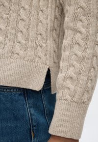 Gros plan sur la manche et l’ourlet avec une fente latérale d’un pull beige en maille torsadée porté sur un jean bleu.