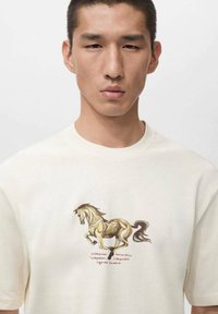 Kermanvärinen puuvillainen t-paita, jossa on ruskeankeltaisen hevosen kuva ja teksti alla. Lyhyet hihat ja klassinen pyöreä kaula-aukko.