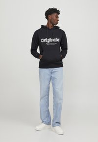 Sudadera con capucha negra que lleva el texto blanco "Originals", bolsillo frontal y cordones. Combinada con jeans de un azul claro y zapatillas blancas.