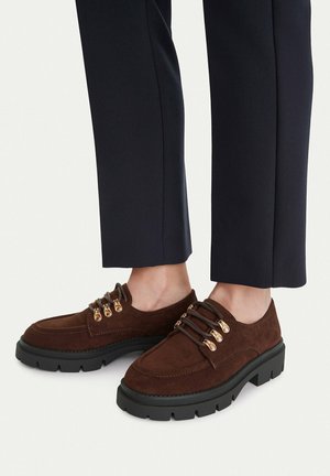 Braune Schnürschuhe aus Wildleder mit dicken schwarzen Sohlen, getragen zu schwarzen, gekürzten Hosen an nackten Füßen.