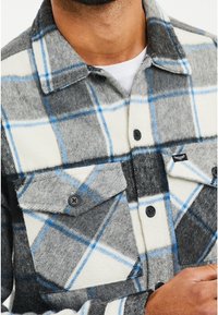 Camicia da flanella a quadri grigi e bianchi con linee blu, dotata di taschini sul petto, bottoni neri e un tessuto morbido e texture.