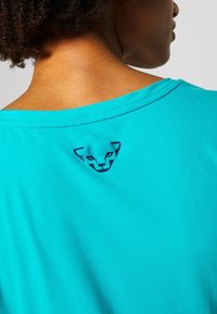 Camiseta deportiva turquesa con un escote amplio, que presenta un logo azul de una cara estilizada cerca del cuello en la parte trasera. Textura suave.