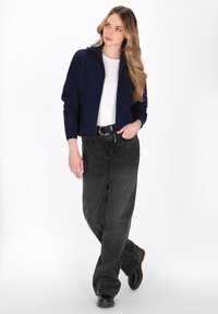 Frau mit langen, welligen Haaren, die einen navy blauen Reißverschluss-Pullover, ein weißes T-Shirt, lockere schwarze Jeans, einen schwarzen Gürtel und schwarze Stiefel trägt, steht mit einer Hand in der Tasche.