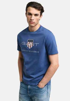 Jonge man draagt een blauw GANT T-shirt met een wapenschild uit 1949 en de tekst "American Sportswear", gecombineerd met lichtblauwe spijkerbroek, handen in de zakken.