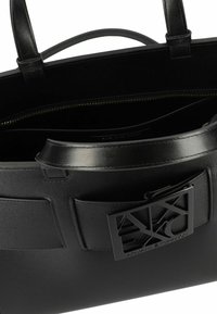 Zwarte leren handtas met een gestructureerd ontwerp, voorzien van een opvallende logo-plaat, twee handvatten bovenaan en een binnenzak met ritssluiting.