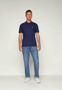 Marineblå polo-shirt lavet af bomuld med en kort krave og et gult logo på brystet. Båret sammen med lyseblå jeans og hvide sneakers.