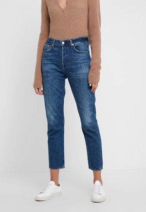 Personne portant un jean bleu taille moyenne, des baskets blanches et un pull en tricot marron à manches longues avec un profond col en V.