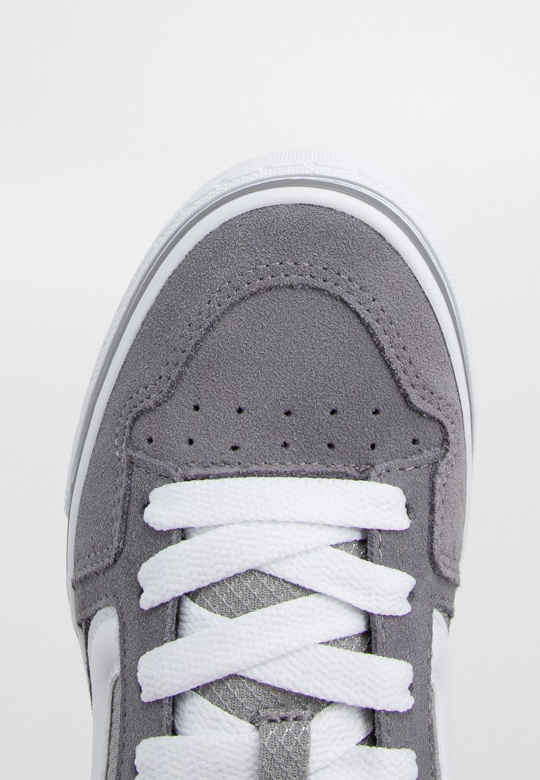 Zapatilla de ante gris con cordones blancos, suela de goma y puntera perforada para transpirabilidad. Presenta un acento de logo blanco suave en el costado.