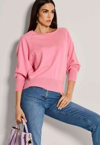 Helles rosa Sweatshirt mit weitem Ausschnitt und gerippten Bündchen, kombiniert mit blauen Jeans. Das Modell hält eine lavendelfarbene Handtasche mit goldenen Hardwareakzenten.