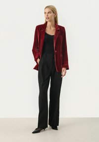 Blazer di velluto rosso con rever a punta, chiusura a singolo bottone e taschino sul petto, abbinato a pantaloni neri a vita alta e tacchi a punta.