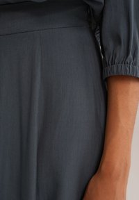 Šedá tkanina s hladkou strukturou, která se vyznačuje volným střihem a nařasenými manžetami. Jednoduchý design s minimem švových detailů.